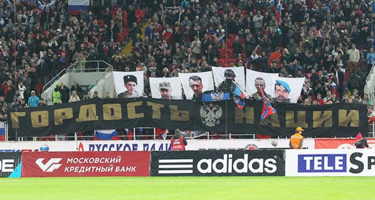 A stand with a black banner reading “ГОРДОСТЬ НАЦИИ”, portraits and Russian flags, the stadium’s advertising boards below.