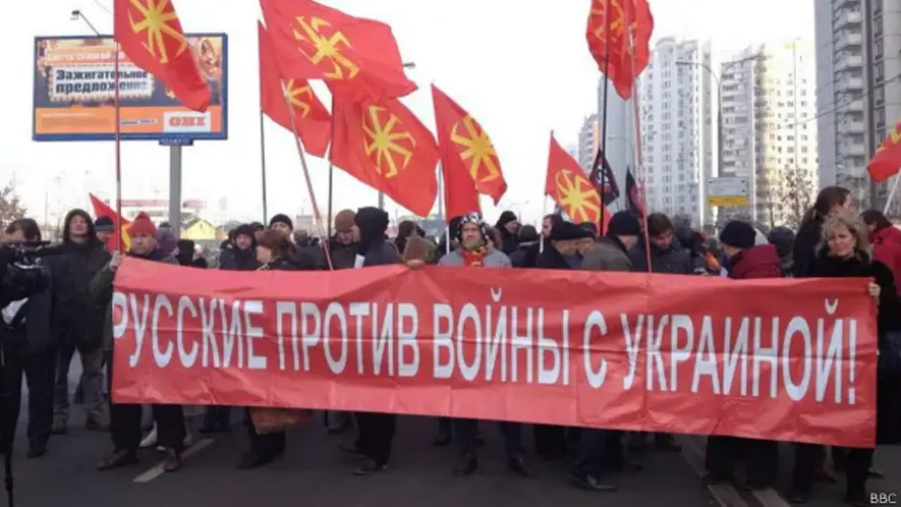 A march under red kolovrat flags, led by a banner reading “РУССКИЕ ПРОТИВ ВОЙНЫ С УКРАИНОЙ!”.