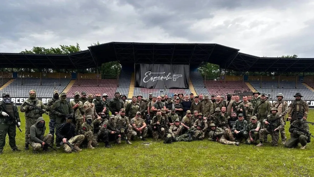 Several dozen armed men in mixed field uniforms pose on a stadium pitch; an “Española” banner hangs on the stand behind them.