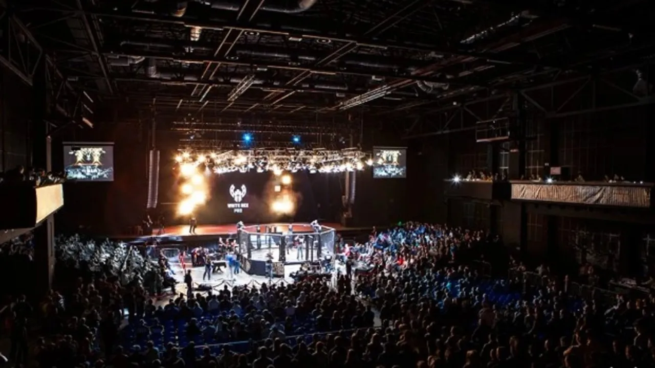 An MMA event in an industrial hall: a lit cage at the centre, the crowd in darkness on the tiers around it.
