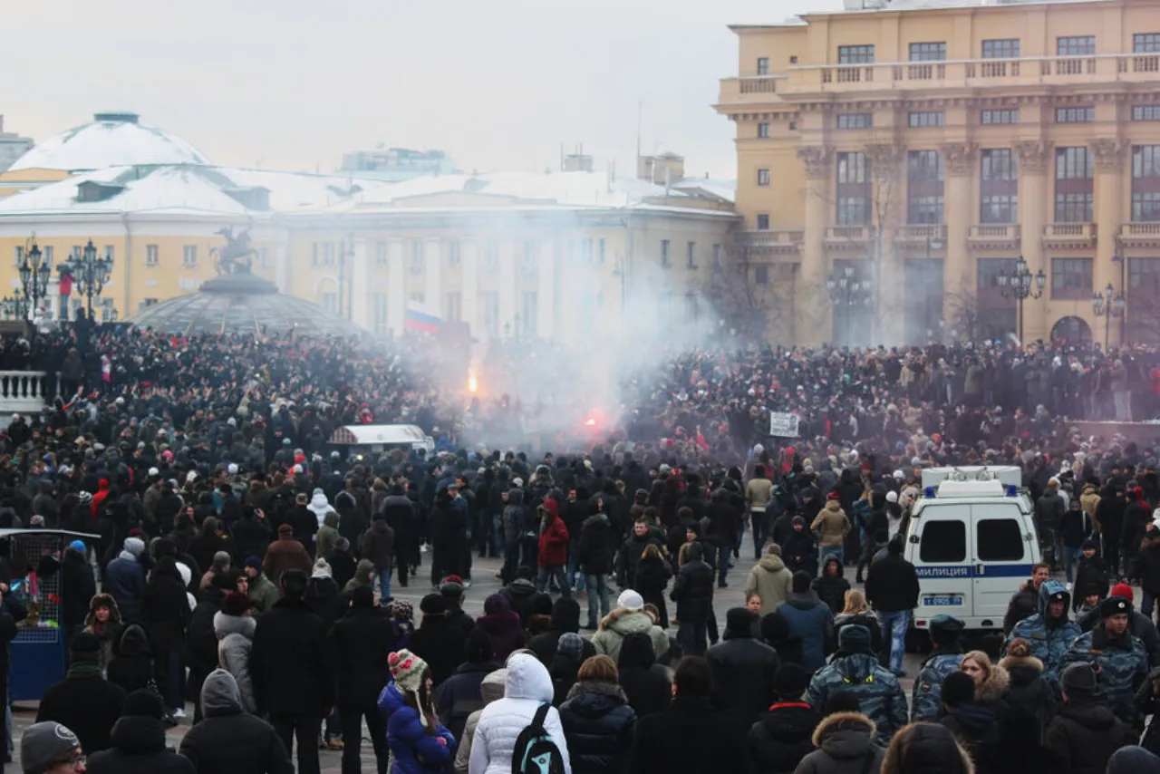 A crowd of several thousand on a Moscow square, flares and smoke overhead, a police van and an OMON cordon in the foreground.