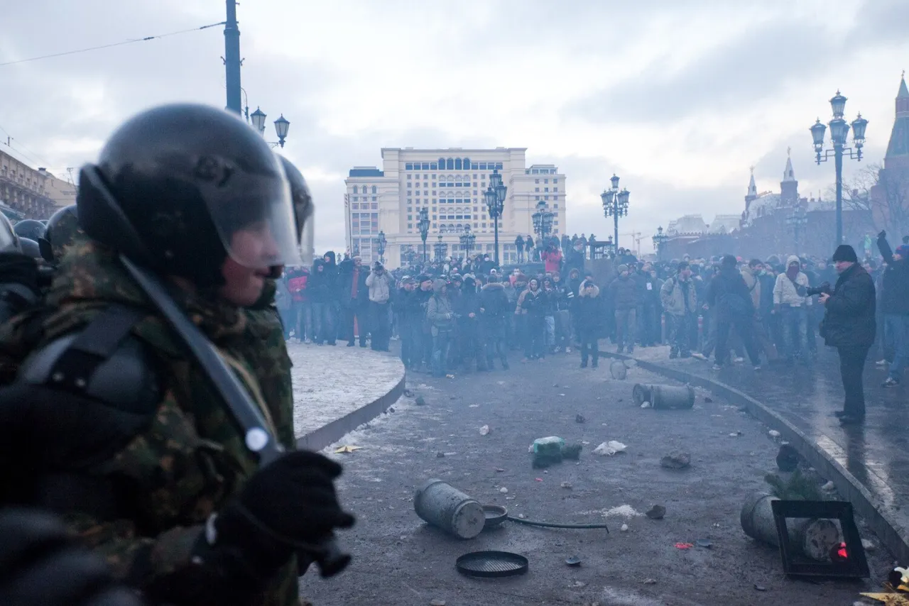An officer in helmet and shield in the foreground, broken paving and the crowd behind him, the Kremlin towers in the distance.