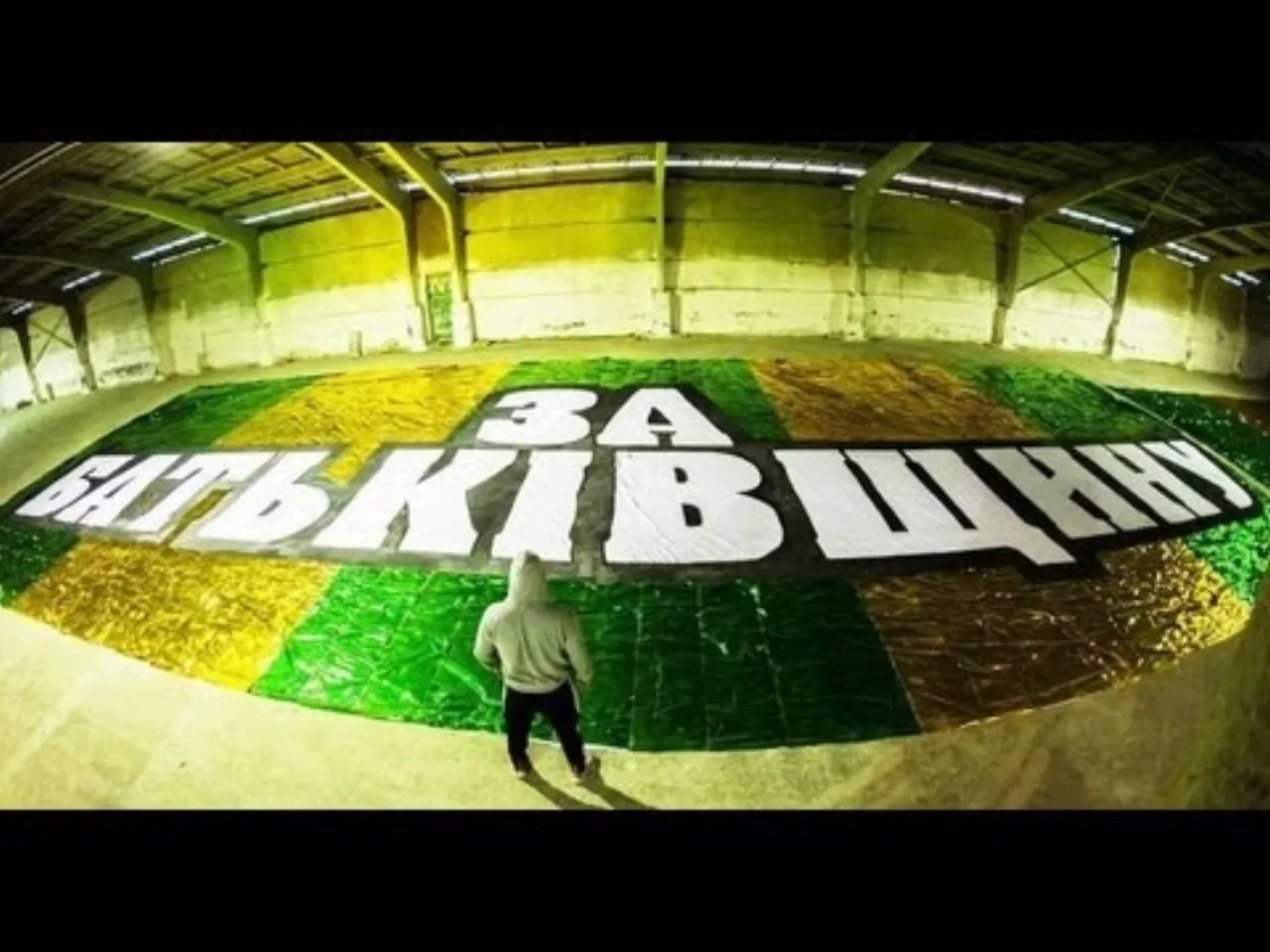 A tifo spread across the pitch, seen from above: a green-and-yellow chequerboard reading “ЗА БАТЬКІВЩИНУ”, a lone figure walking across it.