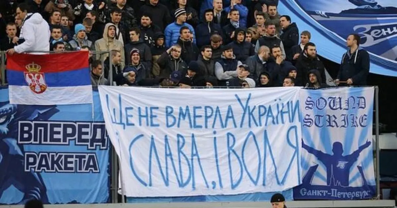 Zenit fans in the stand with banners: a Serbian flag, the words “ВПЕРЕД РАКЕТА”, and a banner carrying a line of the Ukrainian anthem.